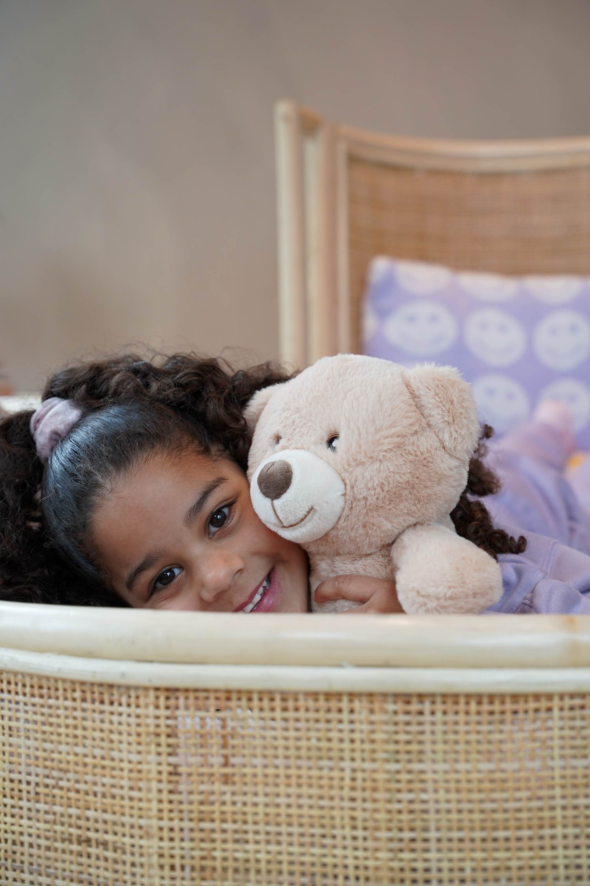 Sfeervolle lifestylefoto van het Rainbow juniorbed van natuurlijk rotan, waarop een kind ontspannen zit. Het handgevlochten hoofdbord en de warme boho uitstraling maken het bed een stijlvolle keuze voor de kinderkamer vanaf circa 5 jaar.