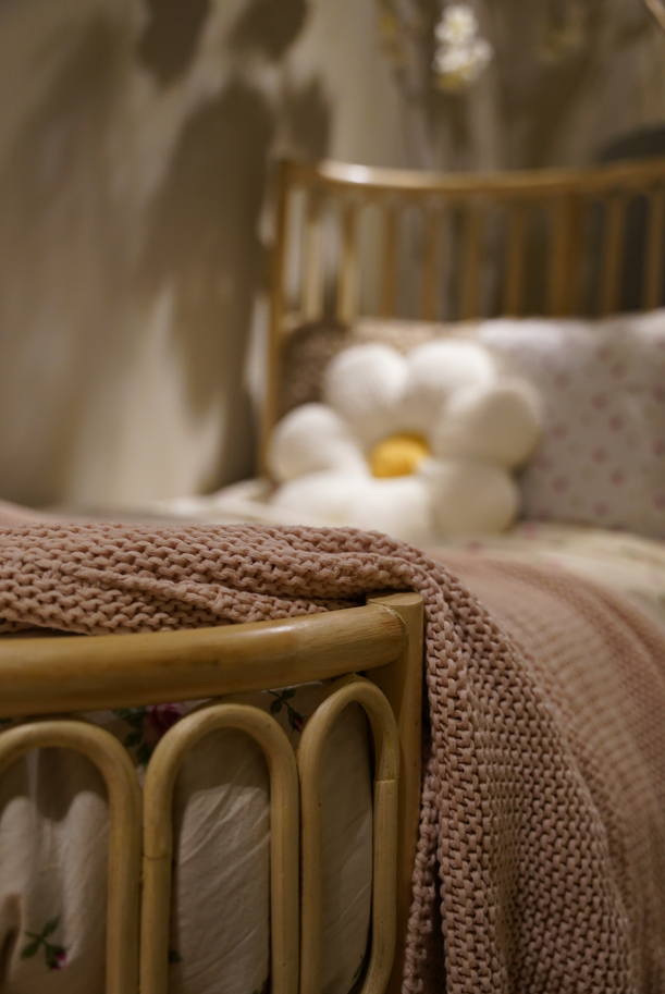Detail van een rotan kinderbed met afgeronde spijlen, gebreide deken en zachte kussens in een sfeervolle kinderkamer