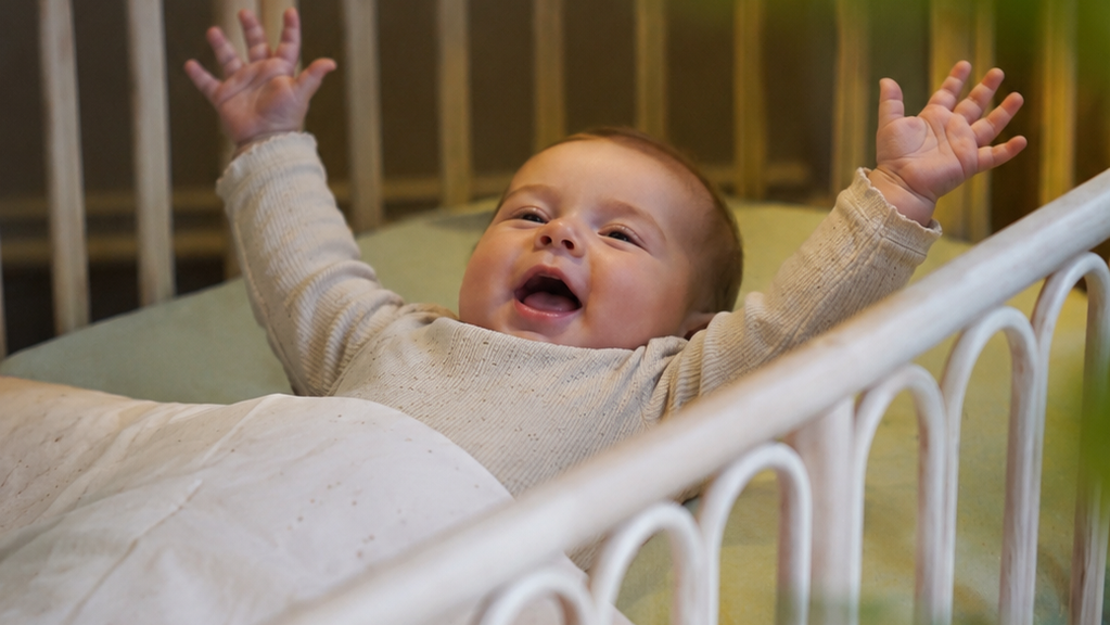 Blije baby in een veilig rotan babybed met afgeronde spijlen en comfortabele matras