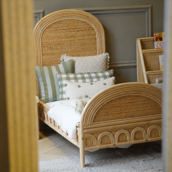 Naturel rotan kinderbed met handgevlochten hoofdbord en afgeronde afwerking in een sfeervolle kinderkamer met kinderboekenkast