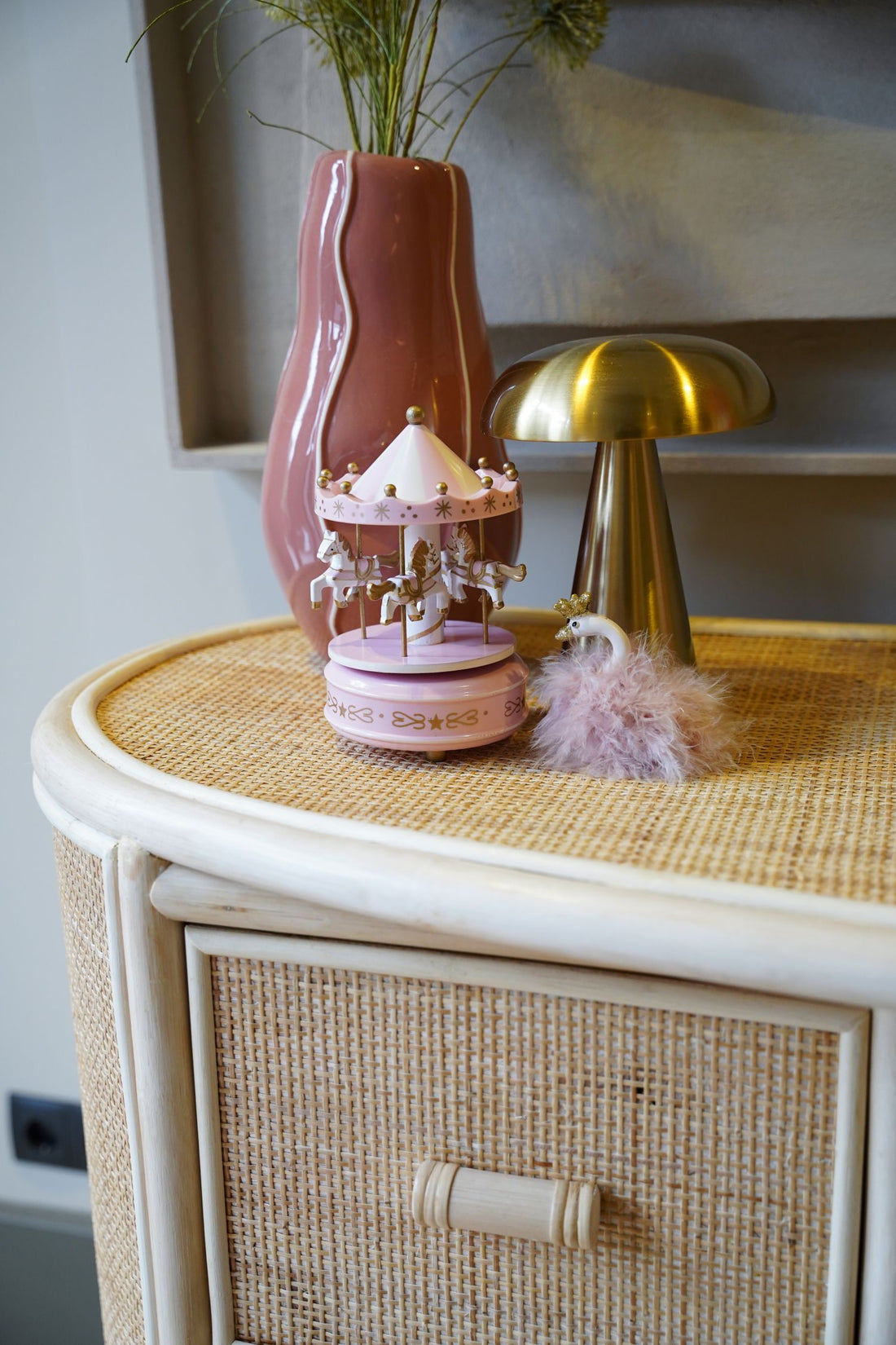 Detailfoto van de Sunny Home Store commode van natuurlijk rotan met handgevlochten afwerking en afgeronde vormen, gestyled met een roze vaas, decoratieve verlichting en een muziekdoos op het ruime bovenblad voor een sfeervolle boho babykamer.