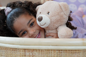 Sfeervolle lifestylefoto van een kind dat knuffelt met een teddybeer in het Rainbow juniorbed van natuurlijk rotan. Het handgemaakte bed met handgevlochten rotan, afgeronde vormen en warme uitstraling creëert een veilige, knusse en geborgen slaapomgeving voor de kinderkamer.