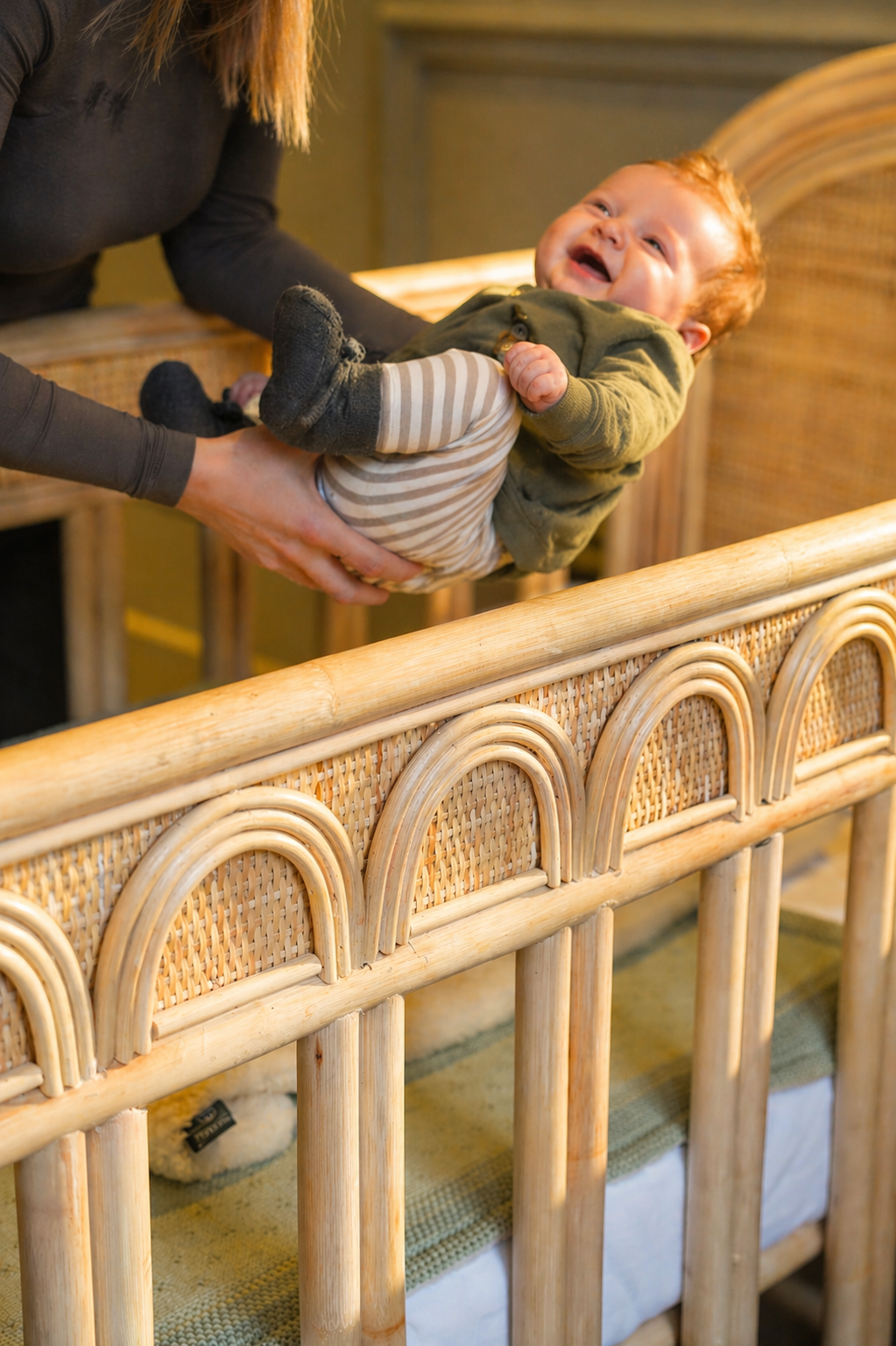 Ouder legt een blije baby veilig in een naturel rotan babybed met handgevlochten details en verstelbare bedbodem