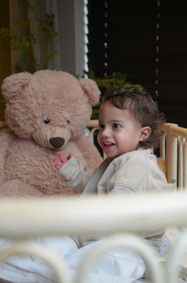 Peuter zit in een naturel rotan peuterbed met knuffelbeer met afgeronde spijlen en een knuffelbeer voor een veilige, sfeervolle kinderkamer